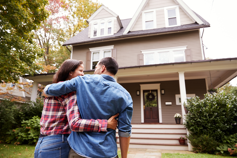 A couple stands arm-in-arm, looking at a two-story house with a porch and steps, surrounded by greenery and trees, on a sunny day.